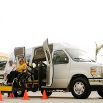 Mulher em cadeira de rodas embarca em uma van acessível por meio de uma plataforma elevatória, com auxílio de um motorista.